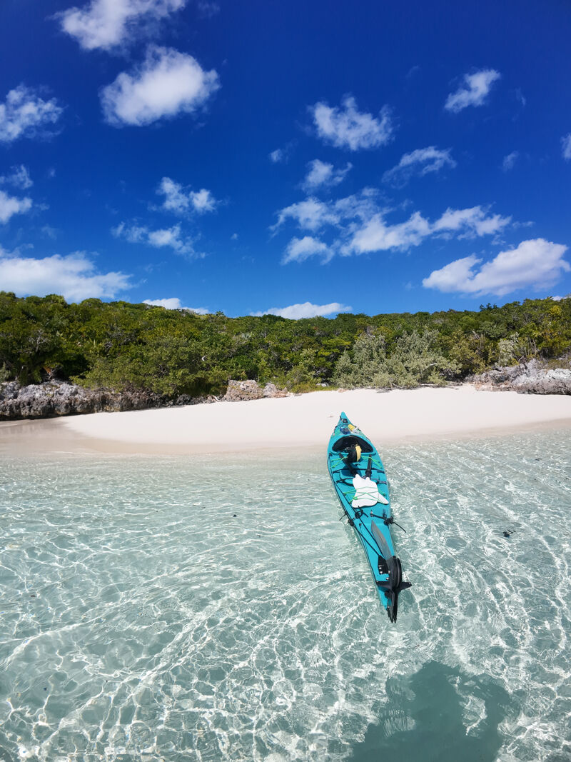 The image showcases a serene coastal scene. A turquoise kayak floats in crystal-clear water, reflecting the bright blue sky dotted with fluffy white clouds. A pristine white sandy beach meets lush green vegetation in the background, creating a picturesque tropical setting. The overall impression is one of tranquility and natural beauty.